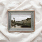 Muted green landscape wall art depicting a peaceful river beneath a rocky hillside